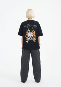Camiseta negra de gran tamaño con un vibrante diseño de calavera rodeado de llamas y el texto "Ed Hardy" con un acabado brillante. Combinada con pantalones gris oscuro.