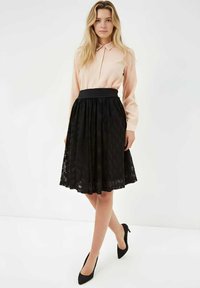 Falda de encaje negro con diseño texturizado, combinada con una blusa de manga larga color rosa claro. Presenta una cintura ajustada y tacones negros.