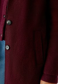 Burgundy wool cardigan with textured fabric, featuring black buttons and a patch pocket. Ribbed cuffs enhance detail and structure.