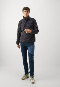 Fleece'owy pulower w ciemnym, wzorzystym materiale z wysokim kołnierzem, zapięciem na guziki z przodu oraz kieszenią na piersi. W zestawie z granatowymi, zwężanymi spodniami i jasnymi butami trekkingowymi.