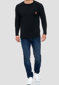 Pull noir en maille avec manches côtelées, encolure ronde et petit patch logo sur la poitrine, associé à un jean en denim bleu et des baskets blanches.