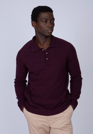 Polo-shirt bordeaux à manches longues avec un col, trois boutons et un ourlet côtelé. Porté avec un pantalon beige clair. Texture lisse.