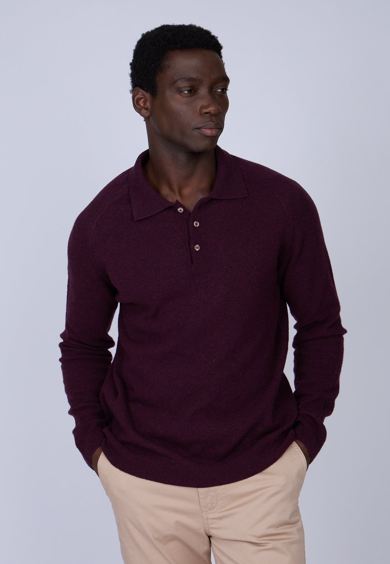 Polo-shirt bordeaux à manches longues avec un col, trois boutons et un ourlet côtelé. Porté avec un pantalon beige clair. Texture lisse.
