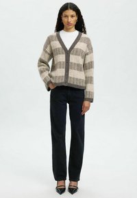 Kreemjas ja pruunis ruuduline kardigan V-kaelusega, nööbist sulgemisega ja ribaga mansetiga, kombineeritud tumedate teksade ja mustade terava ninaga kingadega.