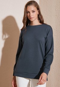 Felpa a maniche lunghe di colore grigio scuro in tessuto morbido, con scollatura rotonda e vestibilità rilassata, caratterizzata da un orlo posteriore leggermente più lungo.