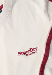 Witte katoenen T-shirt met een rood geborduurd "SuperDry Athletics" logo op de borst en rode accentstrepen langs de schouders.