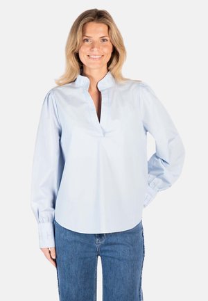 Smilende kvinne står med hånden på hoften, iført lyseblå bluse med langermet mandarinkrage og blå jeans mot hvit bakgrunn.