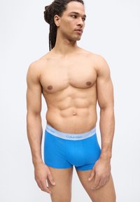 Μυώδης άνδρας με ντραντλοκς που φοράει μπλε μποξεράκια Calvin Klein, κοιτάζοντας προς τα πλάγια με φόντο απλό λευκό.