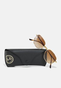 Ray-Ban-solglasögon med guldmetallramar, bruna linser och sköldpaddsmönstrade accenter, vilande på ett svart texturerat fodral med en logotyp.