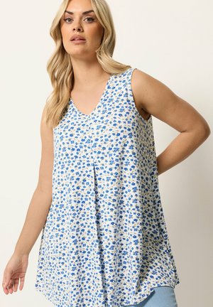 Femme blonde portant un haut blanc sans manches avec un petit imprimé floral bleu, posant avec une main sur la hanche et un fond neutre.