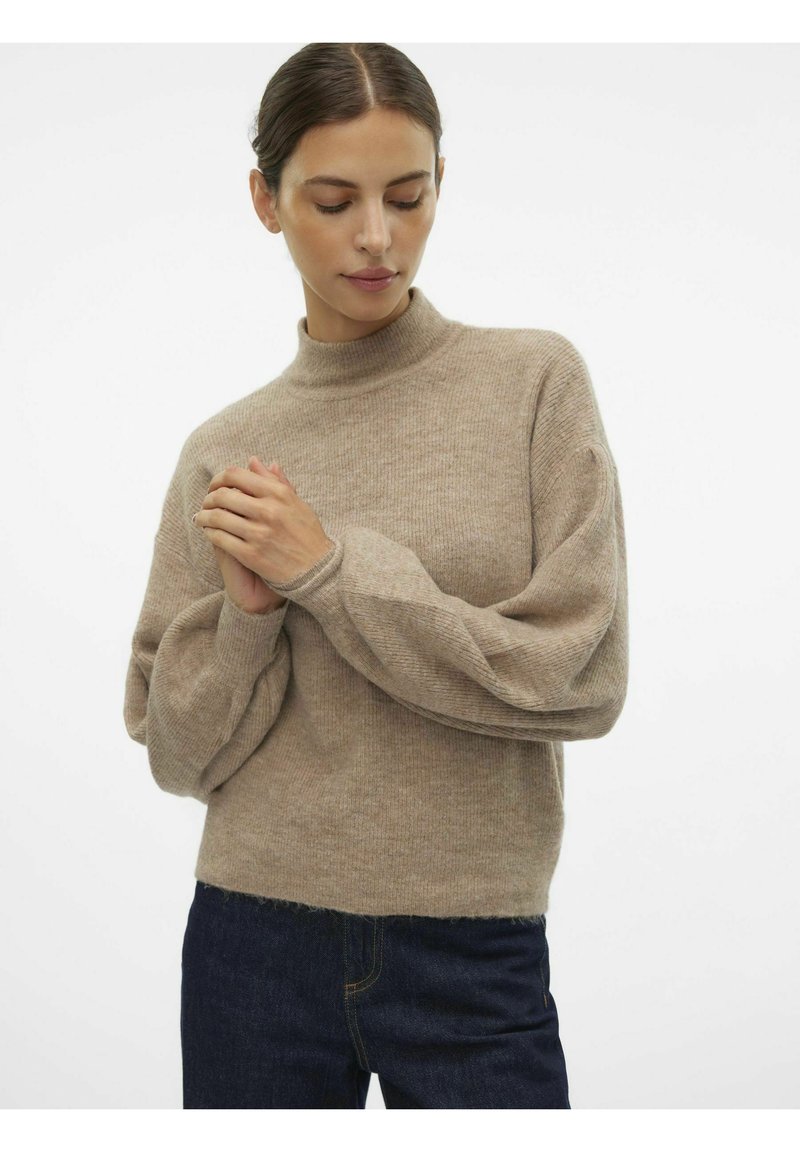 Beige stickad tröja med hög hals och ballongerförsedda ärmar, med ett strukturerat mönster och en avslappnad passform. I kombination med mörka jeans.