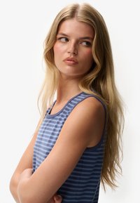 Blonde vrouw met lang haar en roze wangen, gekleed in een blauw gestreepte mouwloze top, kijkt opzij met gekruiste armen.