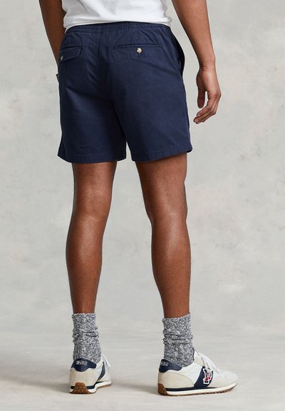 Marinblå shorts med resår i midjan, med två bakfickor med knappstängning. Matchas med vita sneakers och grå strumpor.