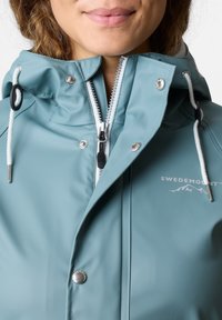 Vattentålig tealjacka med dragkedja på framsidan, dragsko i huvan, silverspännen och en logotyp på bröstområdet. Slät textur, utan mönster.