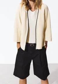 Stickad krämfärgad cardigan med svart kant, trekvartslånga ärmar, bärs över en vit texturerad topp och svarta högmidjade shorts.
