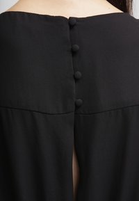 Blusa nera con scollo rotondo, bottoni rivestiti in tessuto sul retro e una leggera apertura per un tocco di originalità. Texture liscia in tutto il capo.