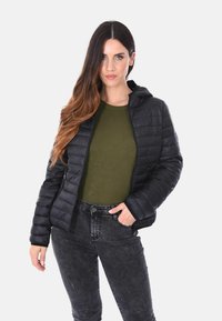 Donna che indossa un giubbotto trapuntato nero sopra un top verde oliva e jeans neri lavati, in piedi con una mano nella tasca.