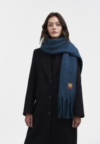 Blå uldscarf med frynser i enderne og et lædermærke med et logo, båret over en sort frakke, på en neutral baggrund.