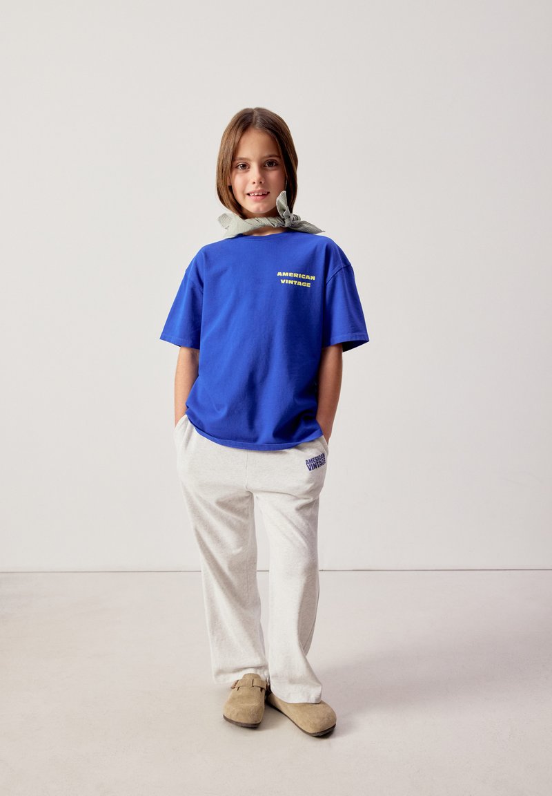 Noor inimene kannab sinist "American Vintage" T-särki, helebeeže värvi pükse, beežikaid libisemisjalanõusid ja kaelas seotud salliga, seisab lõdvestunult.