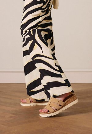 Sandales beiges avec des bretelles croisées, tige texturée et une semelle plateforme blanche. Associées à un pantalon large à motifs noir et crème.