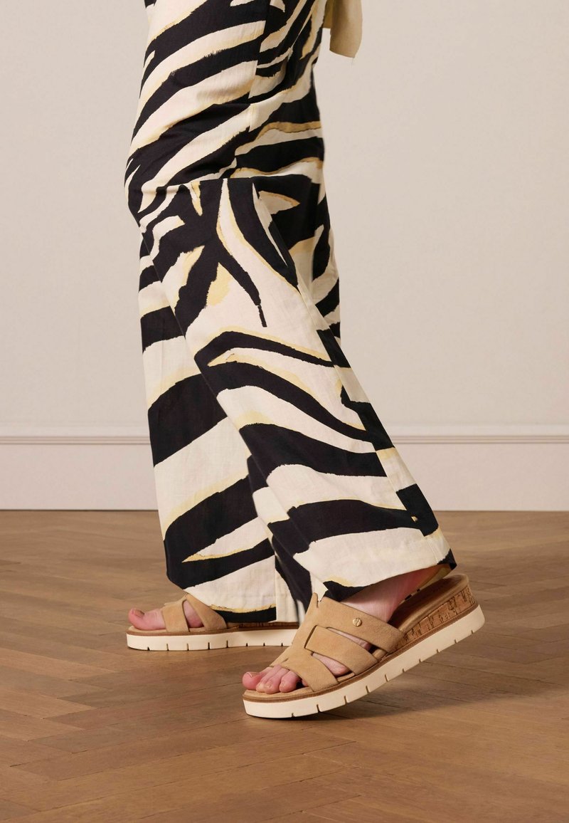 Beige sandalen met kruisbanden, gestructureerde bovenkant en een witte plateauzool. Gecombineerd met zwart en crèmekleurige wijdvallende broeken met een patroon.