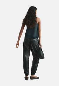 Pantaloni jogger neri in pelle, aderenti alle caviglie, abbinati a un top senza maniche scuro. Tiene una borsa nera con texture, con scarpe basse marroni a pois.