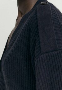Cardigan côtelé bleu marine avec un col en V, présentant un motif de tricot texturé et un accent en bouton sur l'épaule.