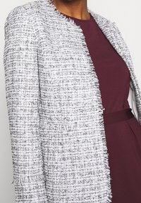 Veste en tweed texturée blanche et grise avec bords effilochés, design à l'avant ouvert, associée à une robe bordeaux en dessous.