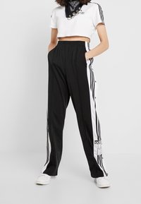 Svarta träningsbyxor med vita sidostripes och tryckknappar, elastisk midja, kombinerat med en kortvit topp och en svart bandana runt halsen.