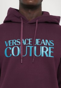 Άτομο που φοράει ένα μωβ φούτερ με τη γραφή "VERSACE JEANS COUTURE" σε μεγάλες ανοικτό μπλε γράμματα στο στήθος.