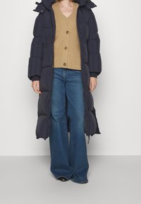 Langer marineblauer Steppmantel mit Kapuze, tailliert und mit Reißverschluss. Kombiniert mit einer beigen Strickjacke und ausgestellten blauen Jeans.