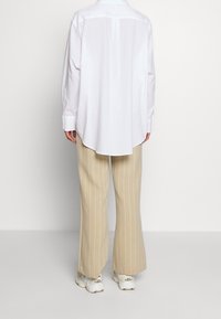 Chemise blanche à boutons avec un ourlet arrondi, associée à un pantalon large beige à fines rayures. Des baskets sont visibles en bas.