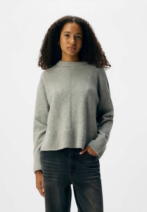 Hellgrauer Pullover mit hohem Kragen, gerippten Bündchen und lockerem Schnitt. Die Textur wirkt weich mit dezenten Nahtdetails.