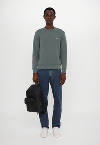Sudadera gris con cuello redondo, combinada con jeans azules. El modelo lleva una mochila negra y usa zapatillas blancas.