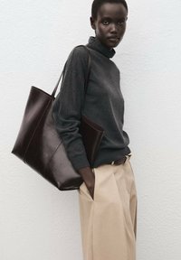 Rjava usnjena torba tote s gladko teksturo, širokim odprtjem in trpežnimi naramnimi trakovi, kombinirana s temnim puloverjem in bež hlačami.