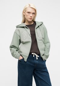 Jeune personne aux cheveux blonds portant un sweat à capuche GAP vert sauge, un t-shirt marron foncé et un jean bleu ample, les mains dans les poches.