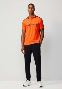 Oranžová polokošile s černými akcenty a potiskem "HACKETT", spárovaná s černými joggingovými kalhotami a bílými teniskami. Jednoduchý design, krátké rukávy.