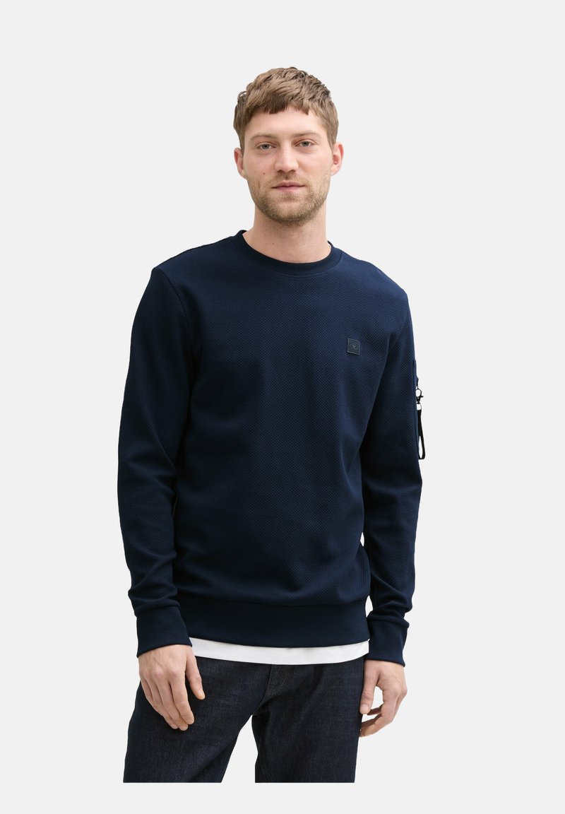 Navy textured sweatshirt with a crew neck, long sleeves, and a zip pocket on the left arm. Layered over a white shirt.