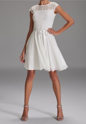 Femme portant une robe blanche en dentelle à manches courtes avec une jupe fluide et des talons hauts à bride cheville, debout avec une main sur la hanche.