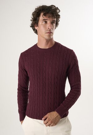 Jersey de punto trenzado en burdeos oscuro, con un patrón texturizado, puños acanalados y un diseño ajustado. Combinado con pantalones de color claro.