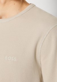 Gros plan sur une personne portant un pull en maille texturée beige avec un col rond et "BOSS" brodé sur la poitrine.