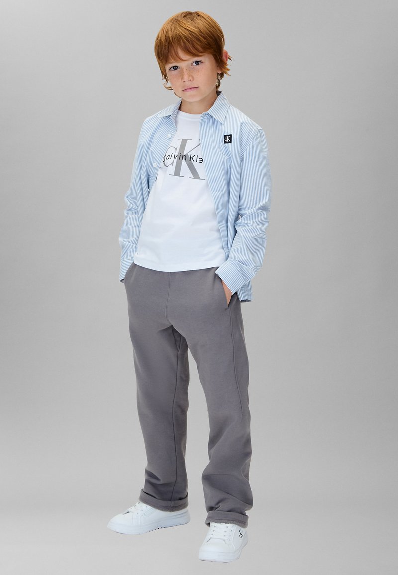 Chemise à rayures bleu clair portée sur un t-shirt blanc avec un logo, associée à un pantalon de survêtement gris et des baskets blanches.