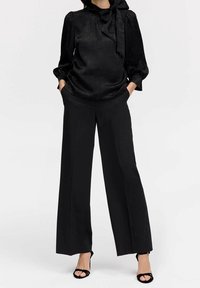 Blusa nera a trama con un fiocco al collo, abbinata a pantaloni neri a gamba larga e tacchi neri con cinturino.