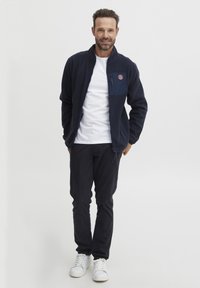 Marineblå fleecejakke med glidelås foran, høy krage og lomme med logo. Brukt med en hvit skjorte og svarte bukser. Hvite sneakers.