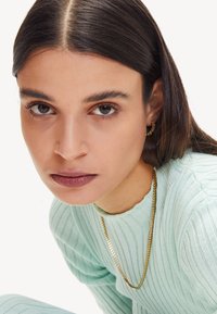 Maglione a coste verde menta con scollatura a girocollo, abbinato a una collana con catena dorata. Caratteristiche principali includono una texture liscia e una leggera lucentezza.