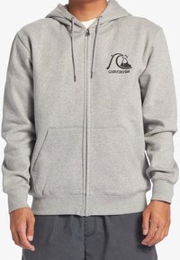 Sudadera gris con cremallera hecha de un tejido suave, con un logo en el lado izquierdo del pecho, bolsillos frontales y capucha ajustable con cordón. Presenta puños y dobladillo acanalados.