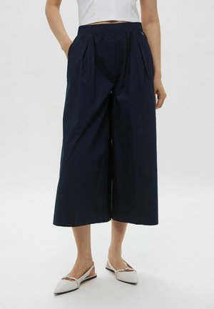 Pantaloni a gamba larga blu navy realizzati in tessuto leggero, con vita elasticizzata, tasche laterali e una superficie liscia. Accentuati da pieghe.