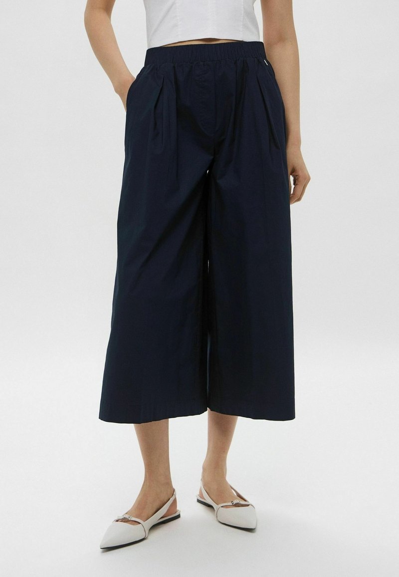 Pantaloni a gamba larga blu navy realizzati in tessuto leggero, con vita elasticizzata, tasche laterali e una superficie liscia. Accentuati da pieghe.