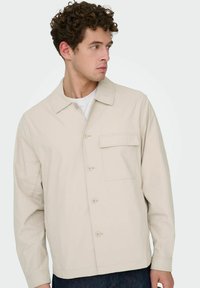 Veste beige à boutons avec col, comprenant une poche poitrine et une texture de tissu lisse. Manches longues et coupe droite.