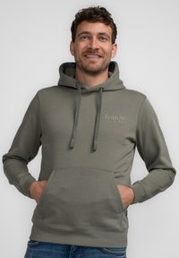 Sudadera verde oliva con bolsillo frontal, capucha con cordón y costuras visibles. Presenta el logo "Petrol Ind. Alaska USA 1989" en verde claro.
